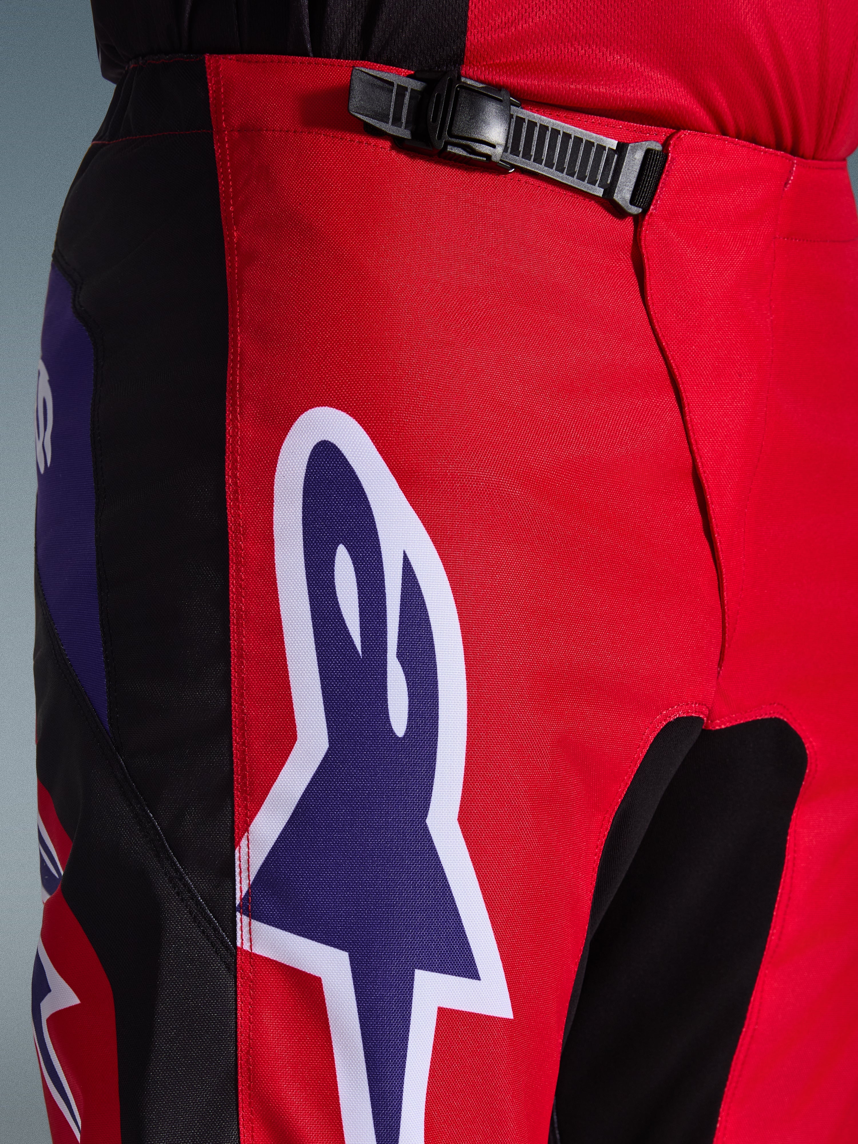 Alpinestars 2026 Fluid Grid bukser, motocrossbukser, rød, sort og lilla, tætbillede af talje- og hofteområdet med en sort skraldelukning og et stort lilla Alpinestars stjernelogo med hvidt omrids, slidstærkt tekstureret materiale til off-road racing.