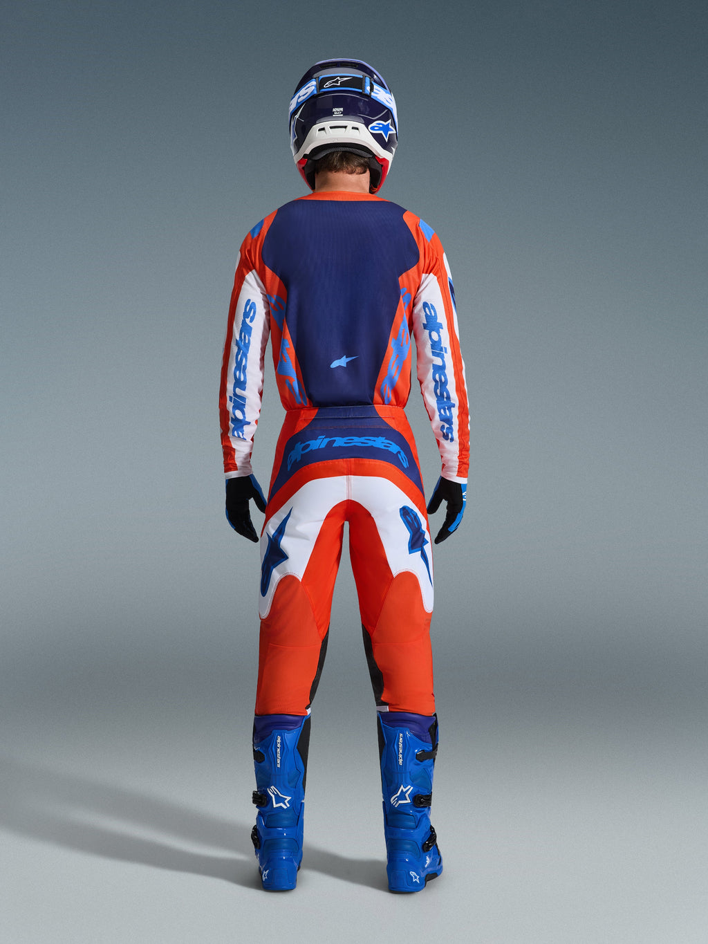 Alpinestars 2026 Fluid Grid bukser, motocross-bukser, orange og UCLA blå, fuld visning bagfra af en kører iført et komplet udstyrssæt med matchende trøje, blå støvler og hjelm, med fremtrædende branding og ergonomiske paneler til terrænkørsel.