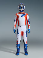 Motocross-kører iført komplet racerudstyr, Alpinestars 2026 Fluid Grid-bukser i Orange og UCLA Blue, hvid jersey med orange ærmer og blå logoer, matchende blå hjelm og goggles, blå støvler og blå handsker, koordineret outfit til off-road kørsel