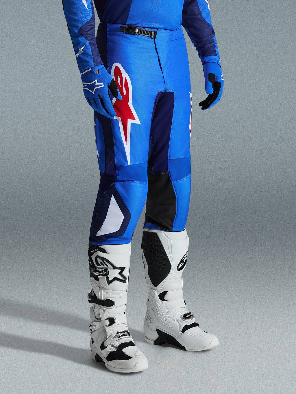 Alpinestars 2026 Fluid Grid bukser, motocross-bukser, blå og rød, båret med hvide motocross-støvler og blå handsker, med et stort rødt stjerne-logo på låret og sorte indvendige knæpaneler, designet til off-road kørsel.
