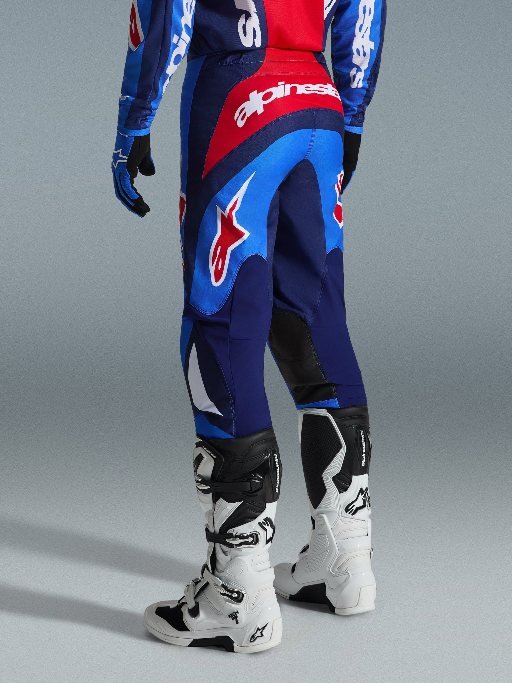 Alpinestars 2026 Fluid Grid bukser, motocrossbukser, blå og rød, set bagfra med markant Alpinestars-branding og ergonomisk design, båret med hvide og sorte Tech 7-støvler og matchende trøje, højtydende udstyr til terrænkørsel