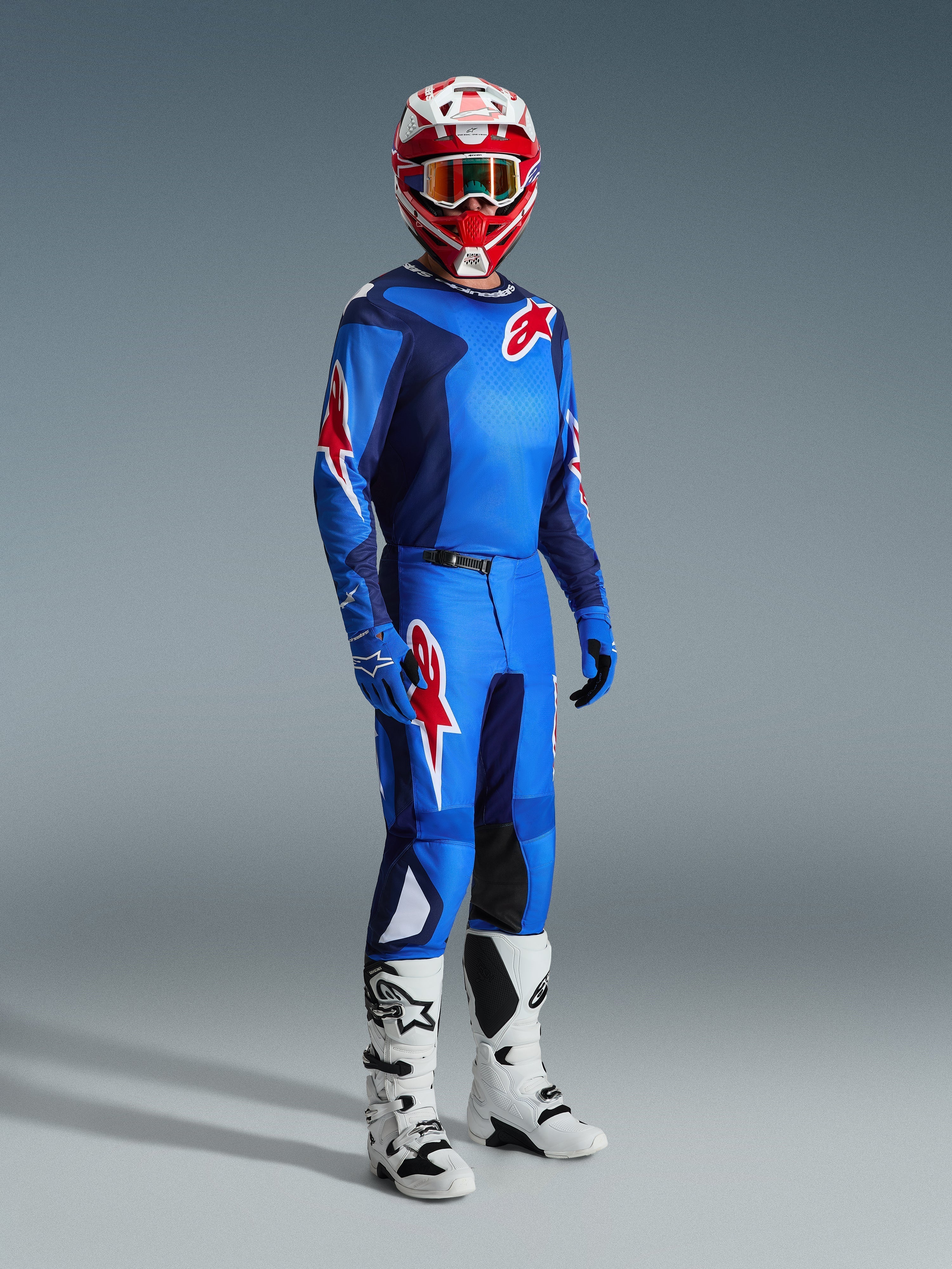 Motocross-kører iført komplet racerudstyr, blå og rød trøje med grid-mønster og Alpinestars-logo, 2026 Fluid Grid-bukser i blå og rød med sorte indvendige knæpaneler, rød og hvid hjelm med crossbriller, hvide og sorte støvler, blå handsker, koordineret sæt til off-road-kørsel