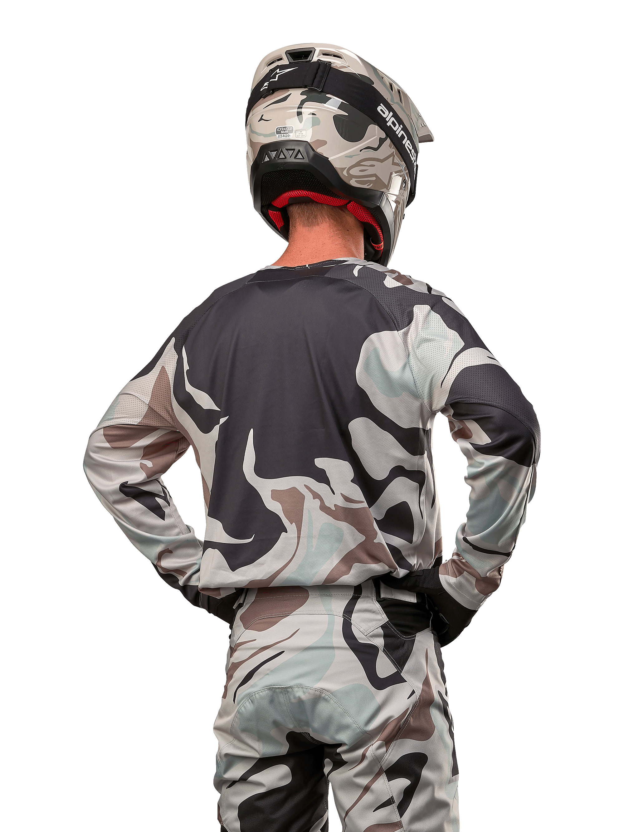 Motocross-kører set bagfra iført Alpinestars 2024 Racer Tactical Jersey i Iron Camo Dust Gray, matchende camouflage-bukser og hjelm, hænderne på hofterne, koordineret off-road racingudstyr.