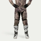 Alpinestars 2024 Racer Tactical bukser, motocross-bukser, militærgrøn camouflage-brun, med store hvide Alpinestars-logoer på lårene og sorte paneler på indersiden af benene, båret med hvide og sorte motocross-støvler, sorte handsker og matchende camouflage-trøje, set forfra.
