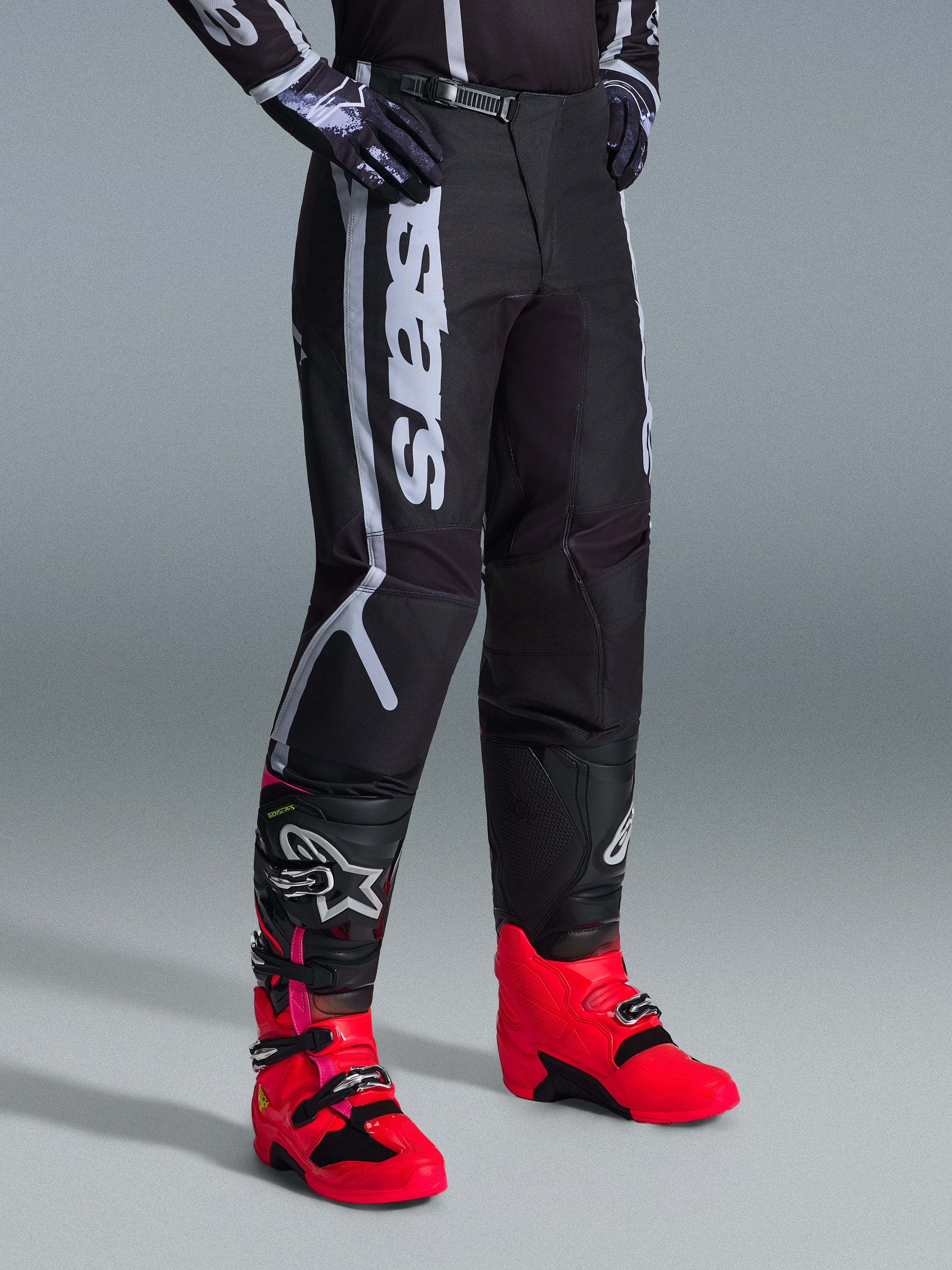 Alpinestars 2026 Fluid Apex bukser, motocross-bukser, sort og grå, båret med livlige pink og røde støvler og matchende sort og hvid trøje, med markant lodret Alpinestars-branding på låret, forstærkede paneler og skraldelukning i taljen for off-road ydeevne.
