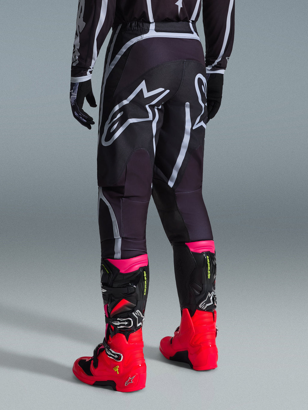 Alpinestars 2026 Fluid Apex bukser, motocrossbukser, sort og grå, set bagfra med store hvide Alpinestars-logoer på sædet, båret med stærkt røde og lyserøde støvler, holdbar 600D polystof-konstruktion til off-road kørsel