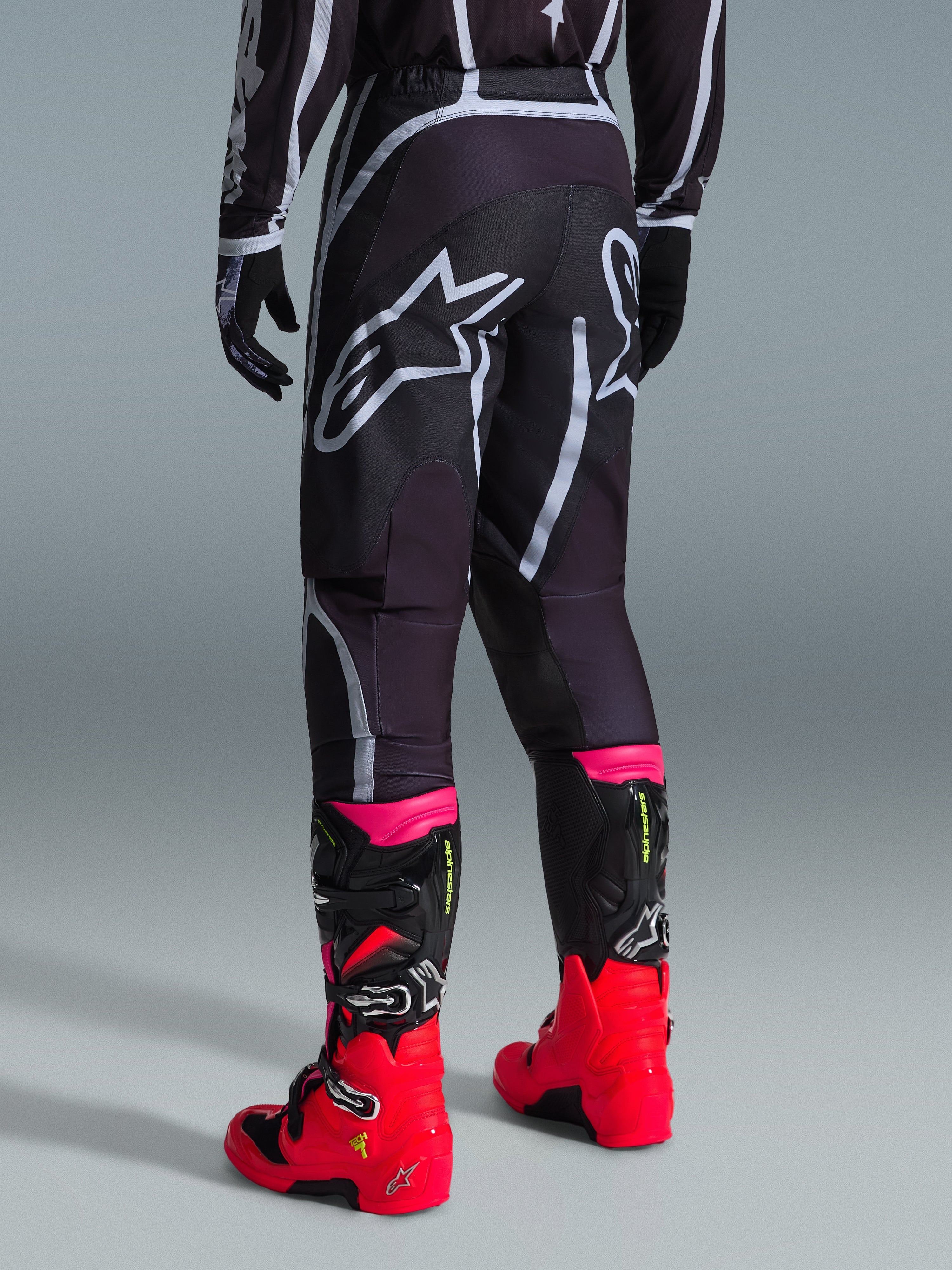 Alpinestars 2026 Fluid Apex bukser, motocrossbukser, sort og grå, set bagfra med store hvide Alpinestars-logoer på sædet, båret med stærkt røde og lyserøde støvler, holdbar 600D polystof-konstruktion til off-road kørsel