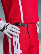 Alpinestars 2026 Fluid Apex bukser, motocross-bukser, rød og hvid, tætbillede af det sorte skraldelukningssystem i taljen, bæres med hvide Alpinestars-handsker og matchende rød trøje, holdbart præstationsudstyr til off-road-kørsel.
