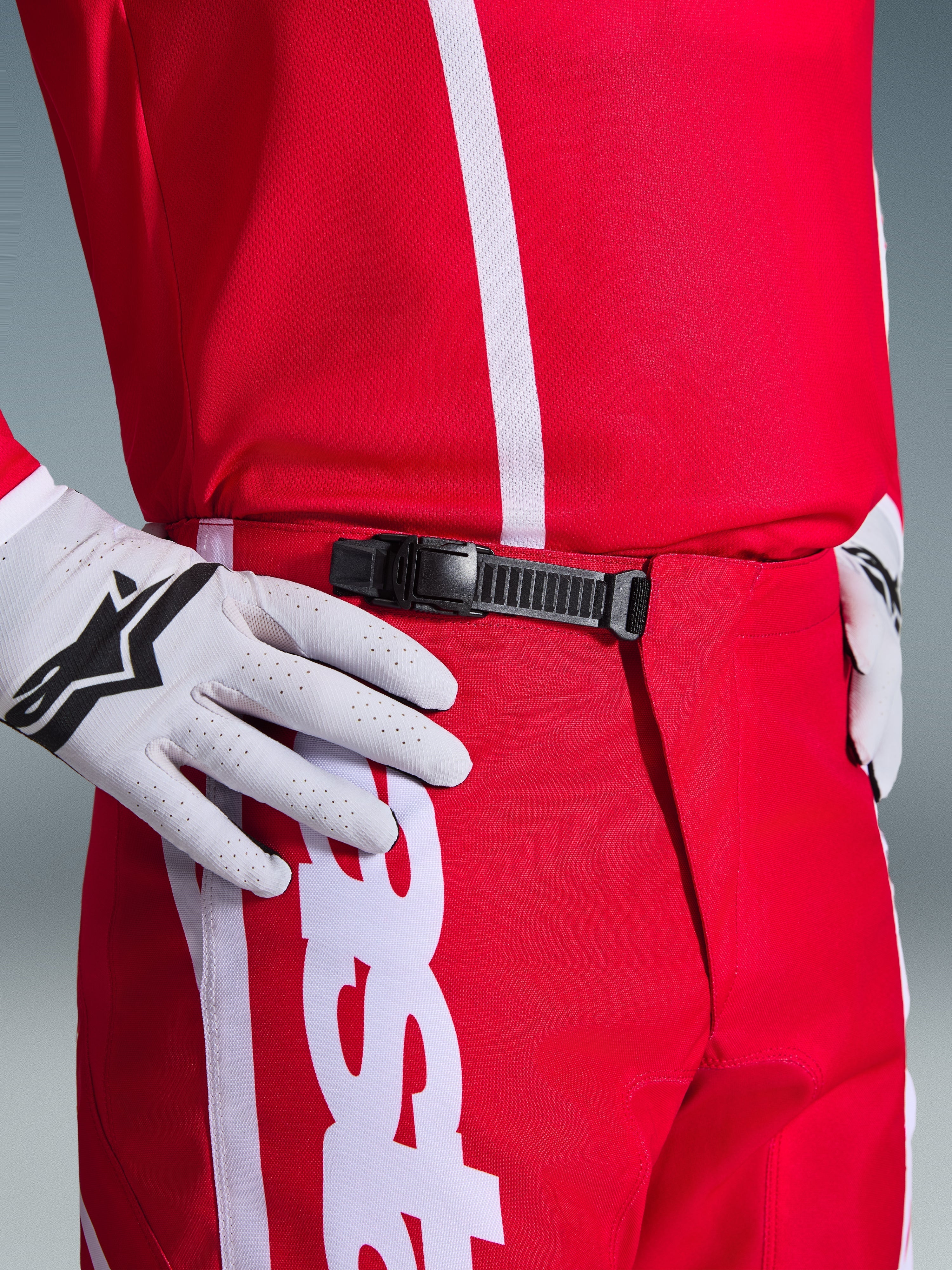 Alpinestars 2026 Fluid Apex bukser, motocross-bukser, rød og hvid, tætbillede af det sorte skraldelukningssystem i taljen, bæres med hvide Alpinestars-handsker og matchende rød trøje, holdbart præstationsudstyr til off-road-kørsel.
