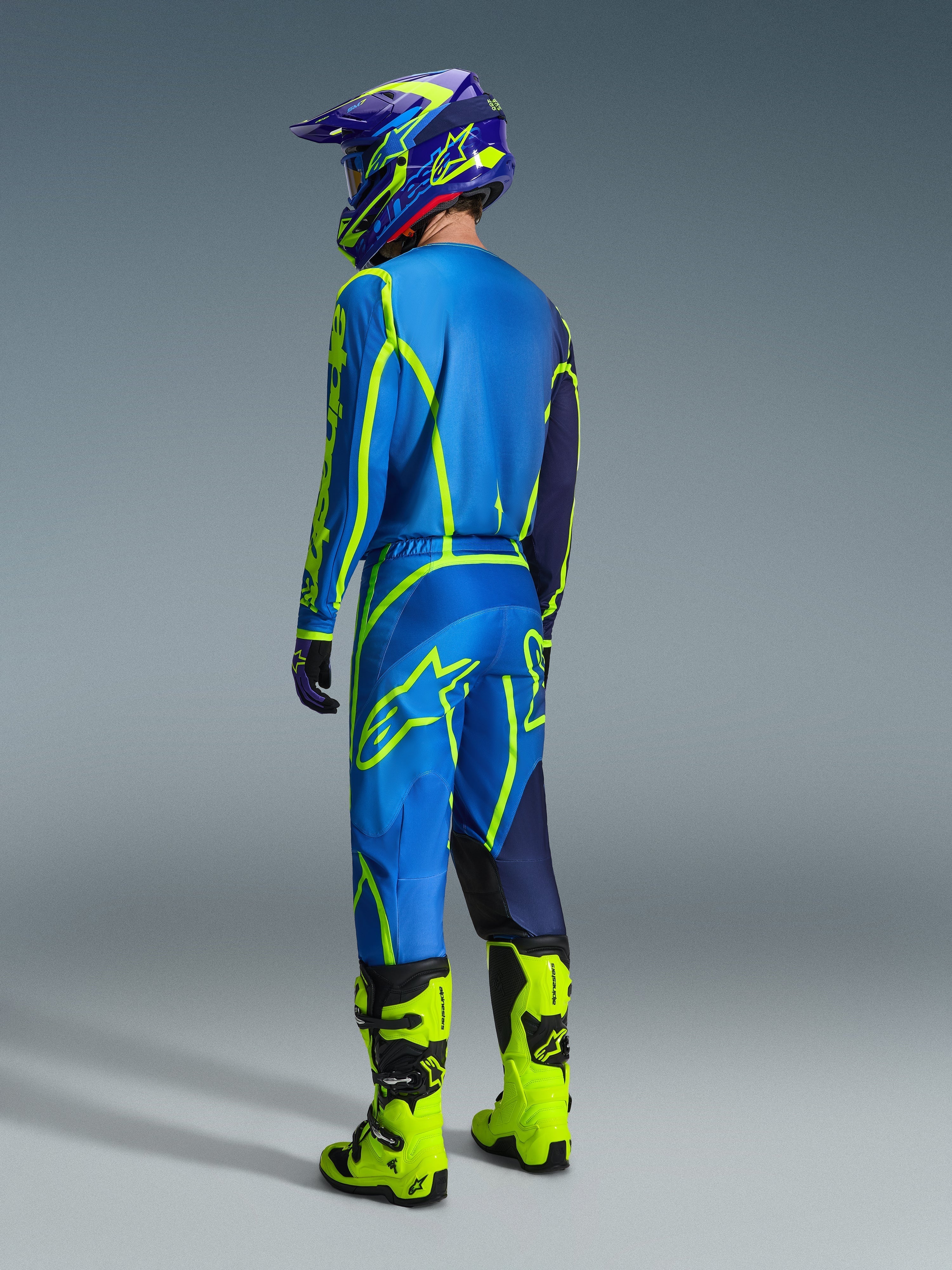 Motocross-kører set bagfra iført 2026 Alpinestars Fluid Apex bukser i blå og neon-gul, matchende blå og gul trøje og hjelm, samt klare neon-gule støvler, koordineret racerudstyr til off-road kørsel.