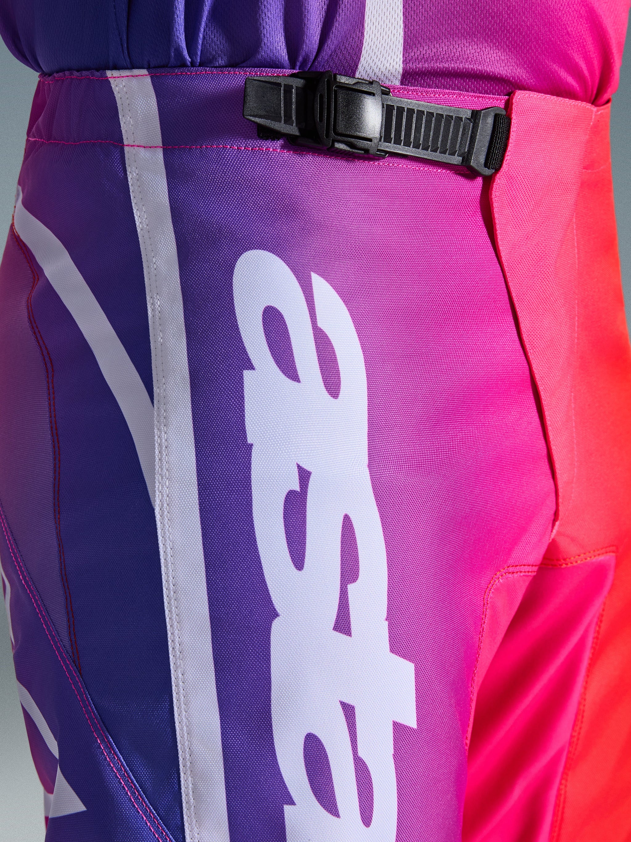 Alpinestars 2026 Fluid Apex bukser, motocross-bukser, multifarvet lilla, pink og rød, tætbillede af talje- og hofteområdet med et sort skraldelukningssystem og stort hvidt lodret Alpinestars-logo, levende gradient-design med holdbar polystof-tekstur