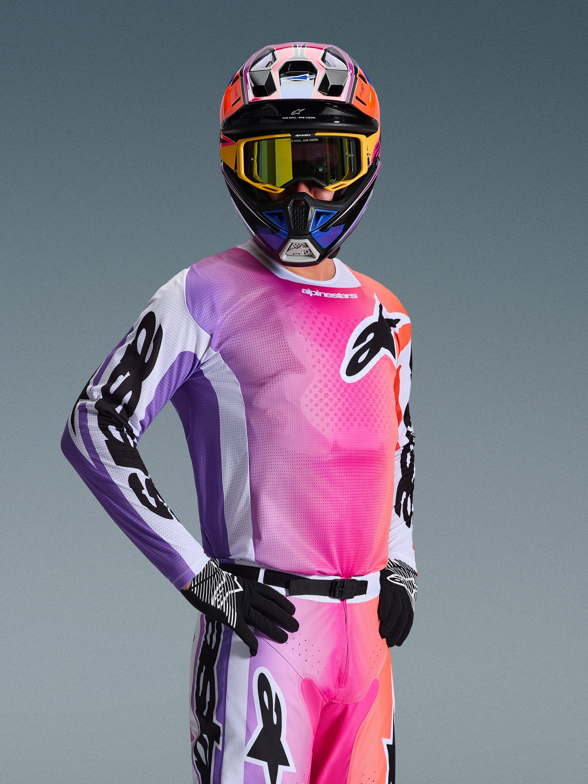 Motocross rider wearing the Alpinestars 2026 Racer Air Portl Jersey, Multicolor pink purple and orange gradient with white accents, matching colorful helmet and yellow goggles, black gloves, professional off-road racing gear