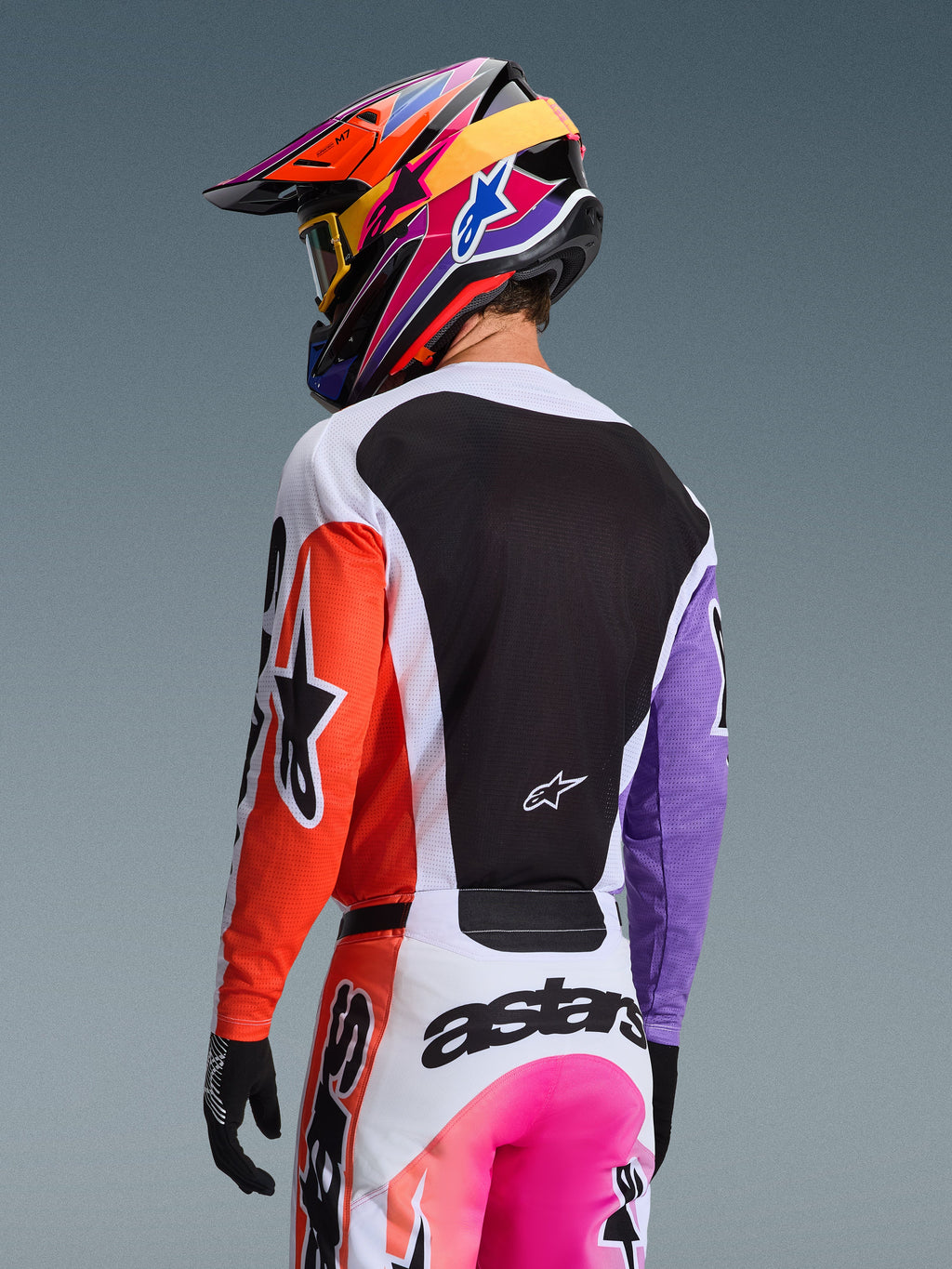 Motocrossrytter set bagfra iført Alpinestars 2026 Racer Air Portl trøje i Multicolor, med et orange højreærme, lilla venstreærme og et stort sort mesh-rygpanel til ventilation, kombineret med matchende motocrossbukser og en farvestrålende flerfarvet hjelm, professionelt off-road racingudstyr.