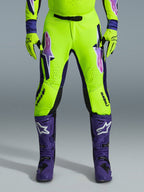 Alpinestars 2026 Supertech Vista bukser, motocross-bukser, fluorescerende gul, lilla og sort, båret med matchende handsker og lilla støvler, med store gradient-logoer på lårene, perforerede zoner til ventilation og et professionelt atletisk design til off-road racing