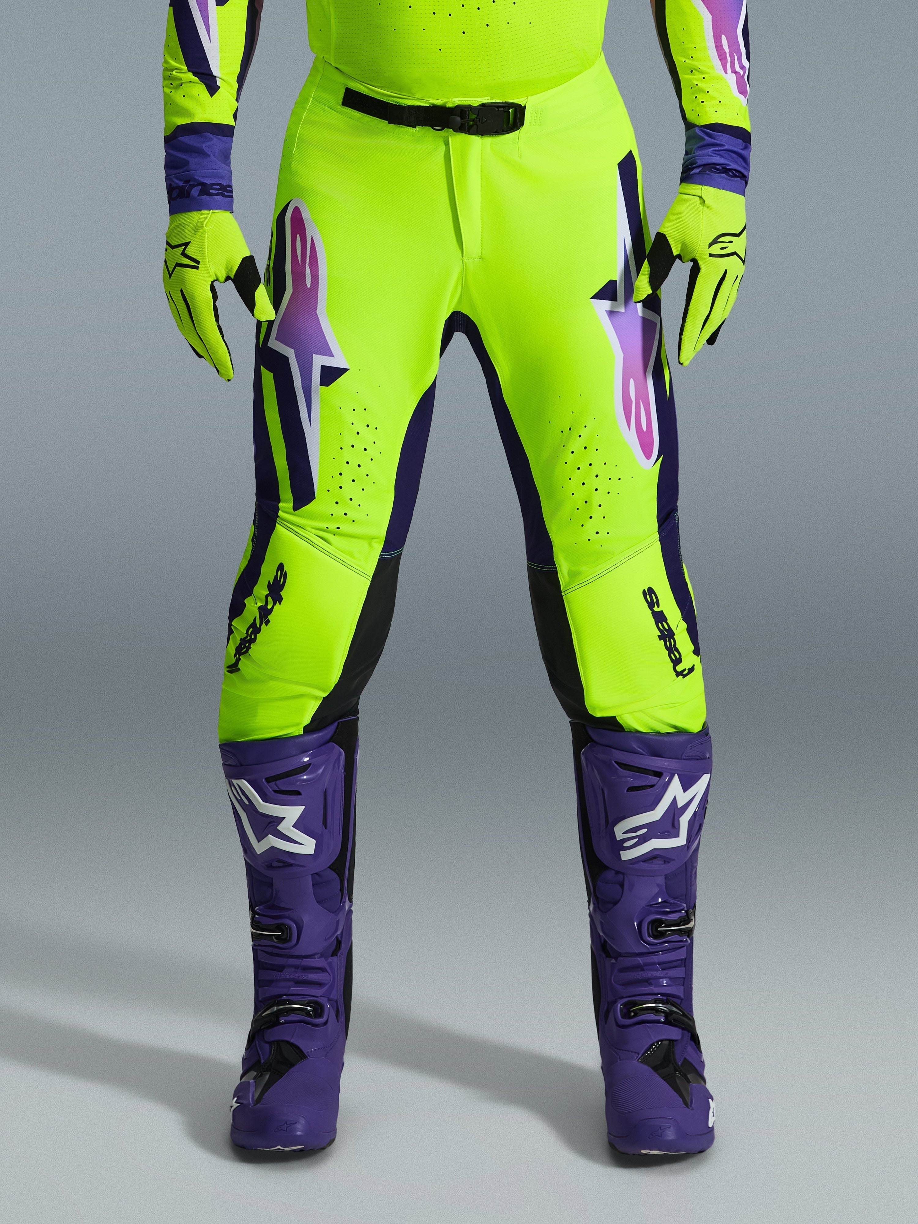 Alpinestars 2026 Supertech Vista bukser, motocross-bukser, fluorescerende gul, lilla og sort, båret med matchende handsker og lilla støvler, med store gradient-logoer på lårene, perforerede zoner til ventilation og et professionelt atletisk design til off-road racing