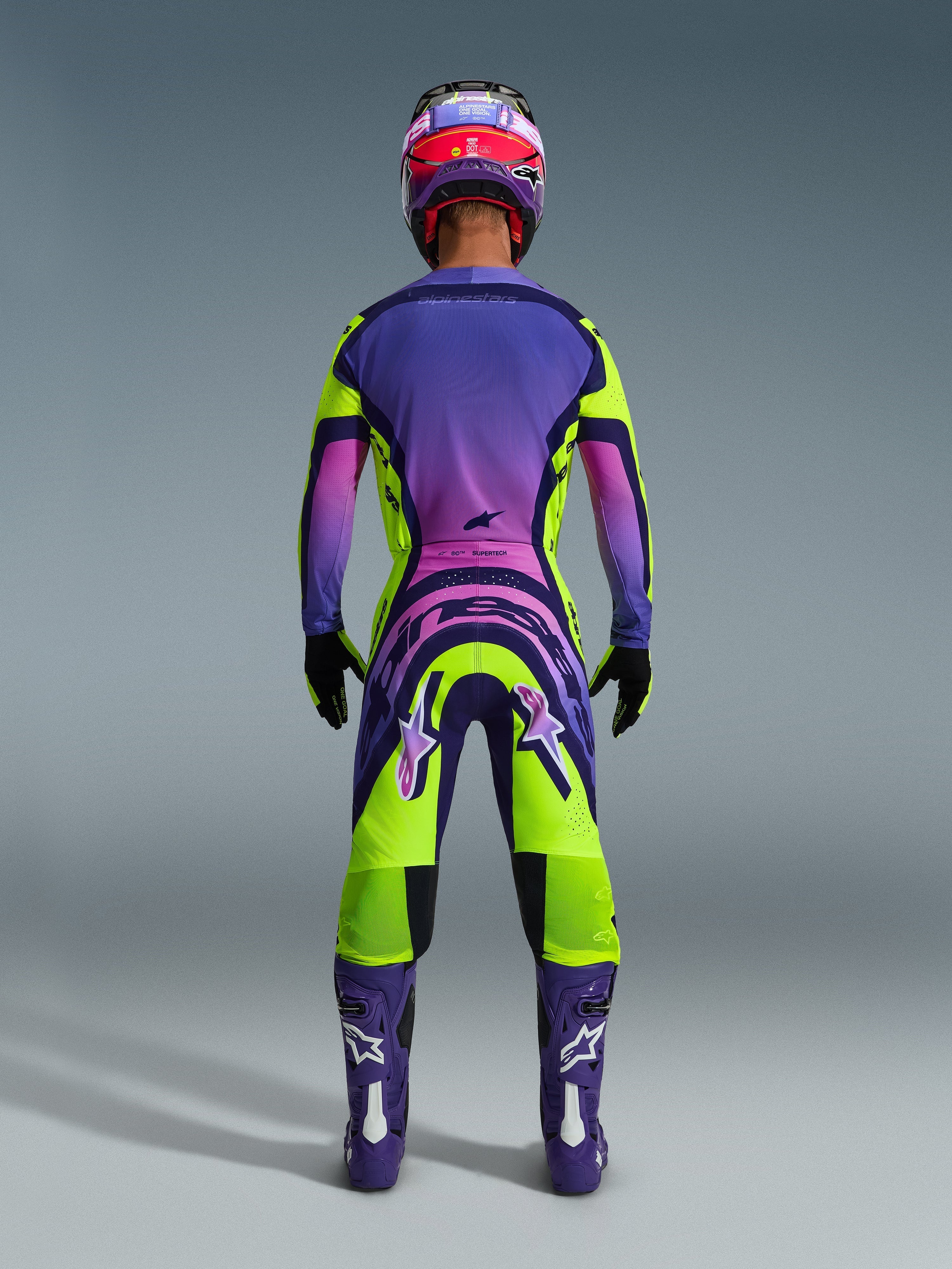 Motocross-kører set bagfra iført 2026 Supertech Vista-trøje og matchende bukser, livligt gult og lilla design med lyserøde gradienter, lilla støvler med hvide Alpinestars-logoer, sorte handsker og en rød og lilla racerhjelm, professionelt racersæt.