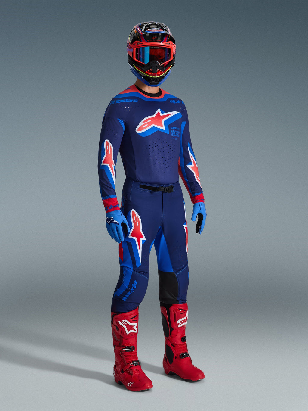 Alpinestars motocrosskører iført 2026 Supertech Vista bukser i Dark Navy Hot Coral Cobalt, matchende langærmet racertrøje, røde motocrossstøvler og koordineret hjelm med røde goggles, helkropsstudiebillede af højtydende off-road udstyr