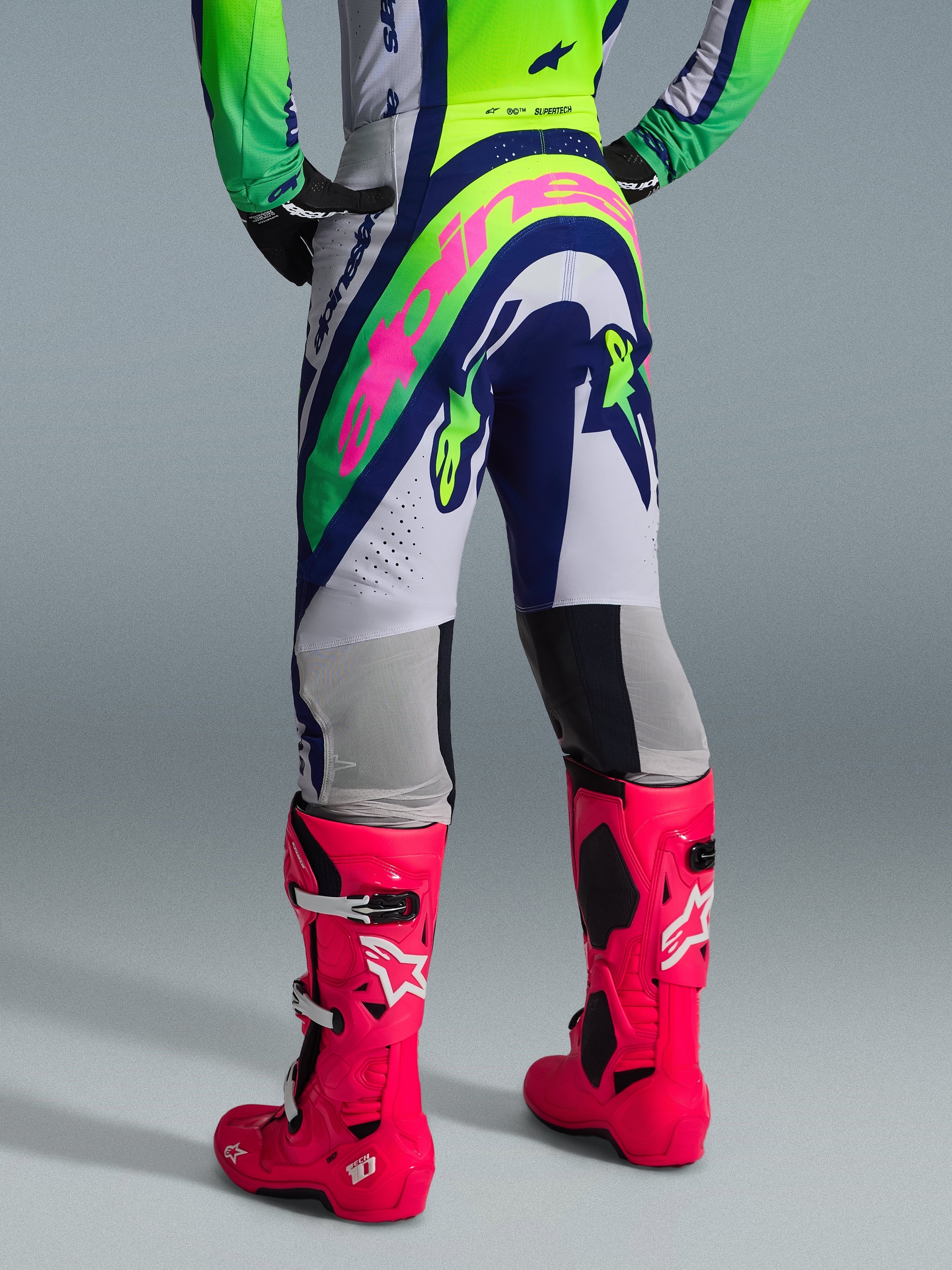 Alpinestars 2026 Supertech Vista bukser, motocrossbukser, lysegrå, neongul og blå, set bagfra med et stort lyserødt Alpinestars-logo, båret med matchende racingtrøje og stærkt lyserøde Tech 10-støvler, professionelt terrænudstyr
