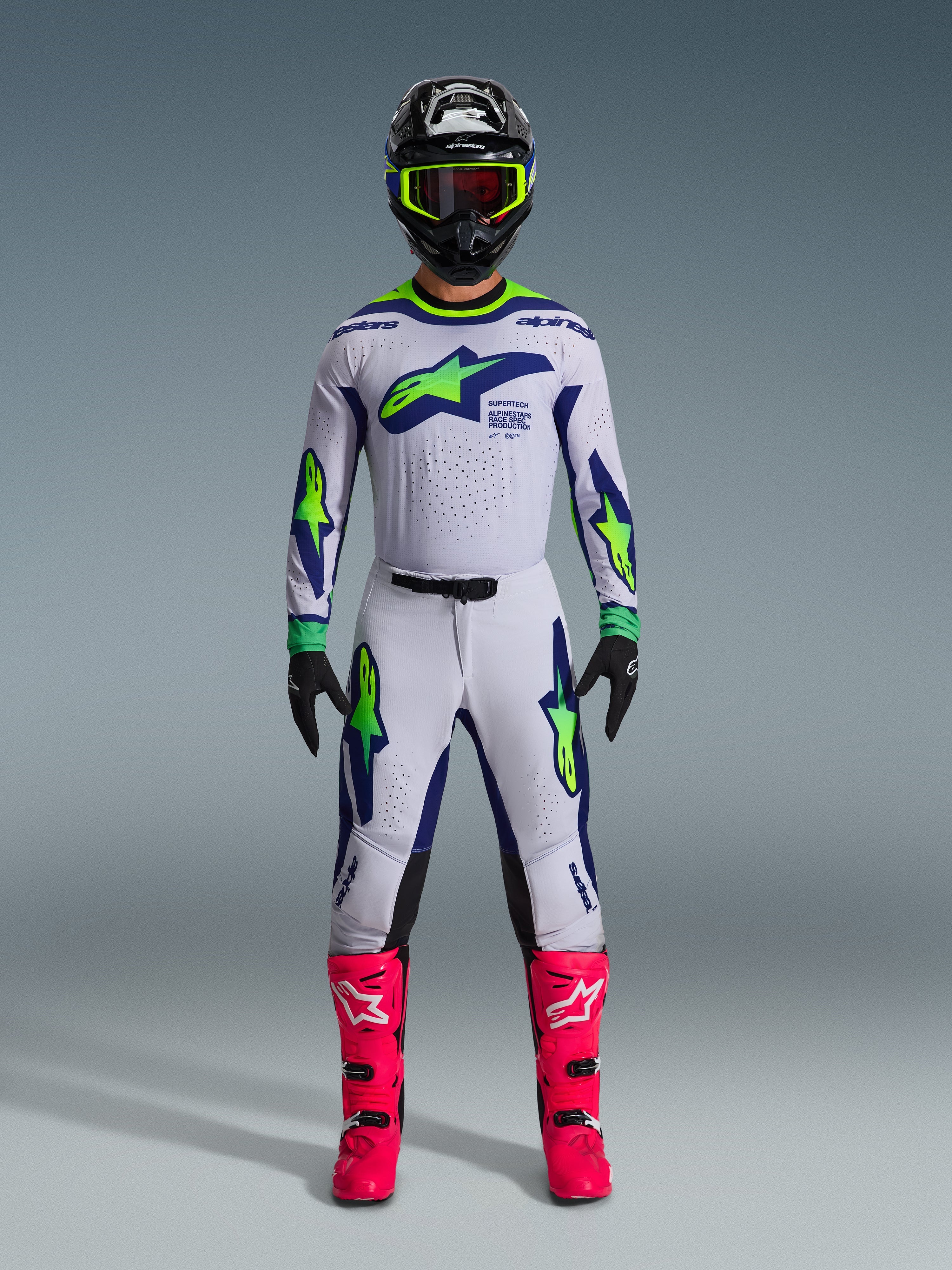 Alpinestars 2026 Supertech Vista Gear Set, motocross-kører iført komplet racing-beklædning, hvid trøje og bukser med blå og neongrønne accenter, sort hjelm med neongule detaljer, stærkt lyserøde støvler, professionelt off-road racing-udstyrssæt