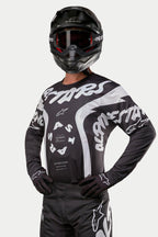 Alpinestars 2024 Racer Hana trøje, motocross-trøje, sort og hvid, med markant hvid Alpinestars-tekstgrafik og logo, båret af en kører med sort hjelm og goggles, højtydende udstyr til off-road racing