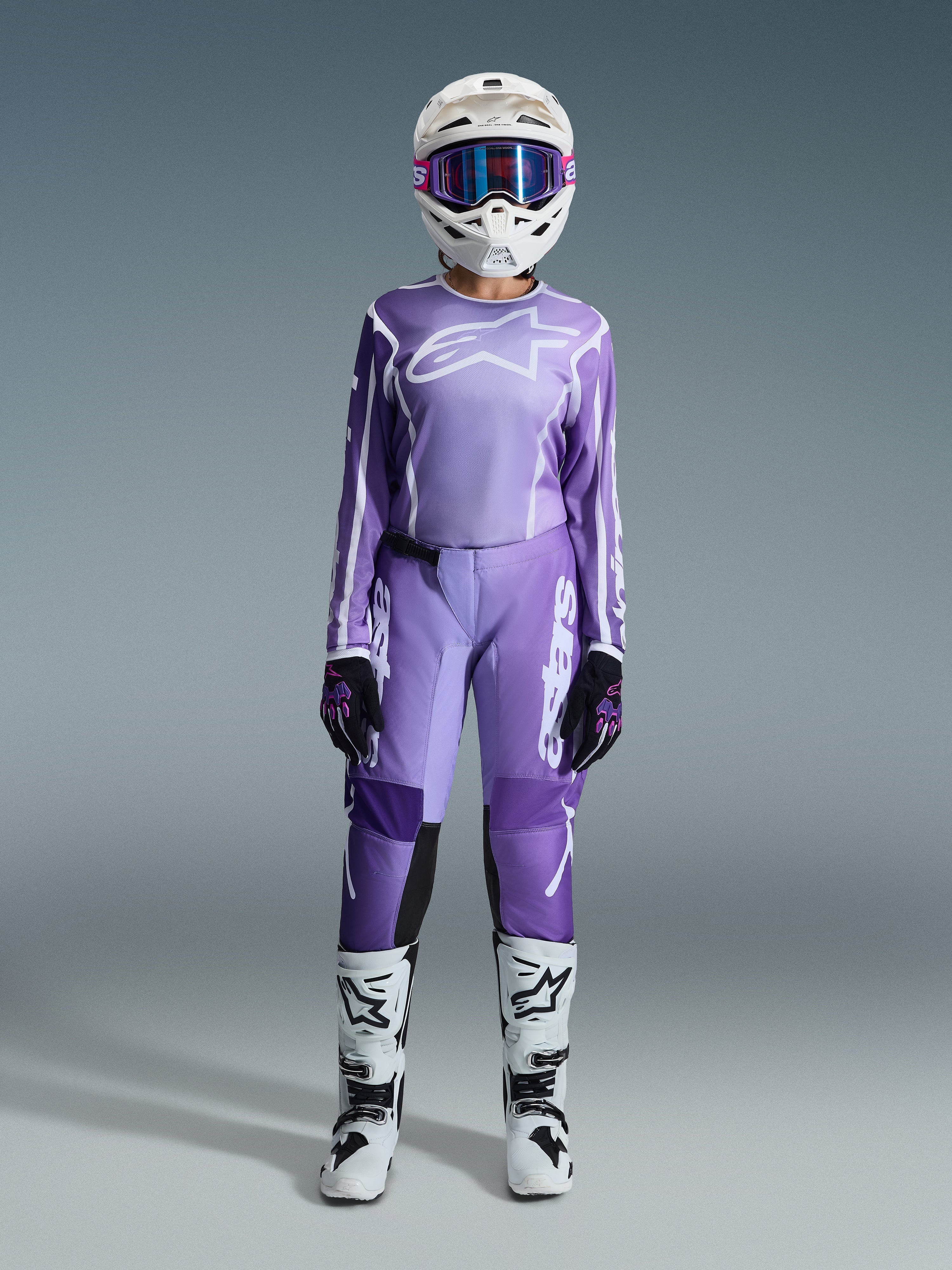 Motocross-rytter i Alpinestars 2026 Stella Fluid Apex-udstyrssæt til kvinder, violet og hvid jersey med matchende bukser, hvid hjelm med lyserøde goggles og hvide og sorte off-road støvler, komplet racersæt til motocross til kvinder.