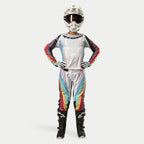 Kvindelig motocross-kører iført Alpinestars Stella 2024 Techstar bukser til damer, Black Multicolor med hvid base samt røde, gule og blå striber, matchende langærmet trøje, hvid hjelm og sorte støvler, komplet racerudstyr til off-road konkurrence