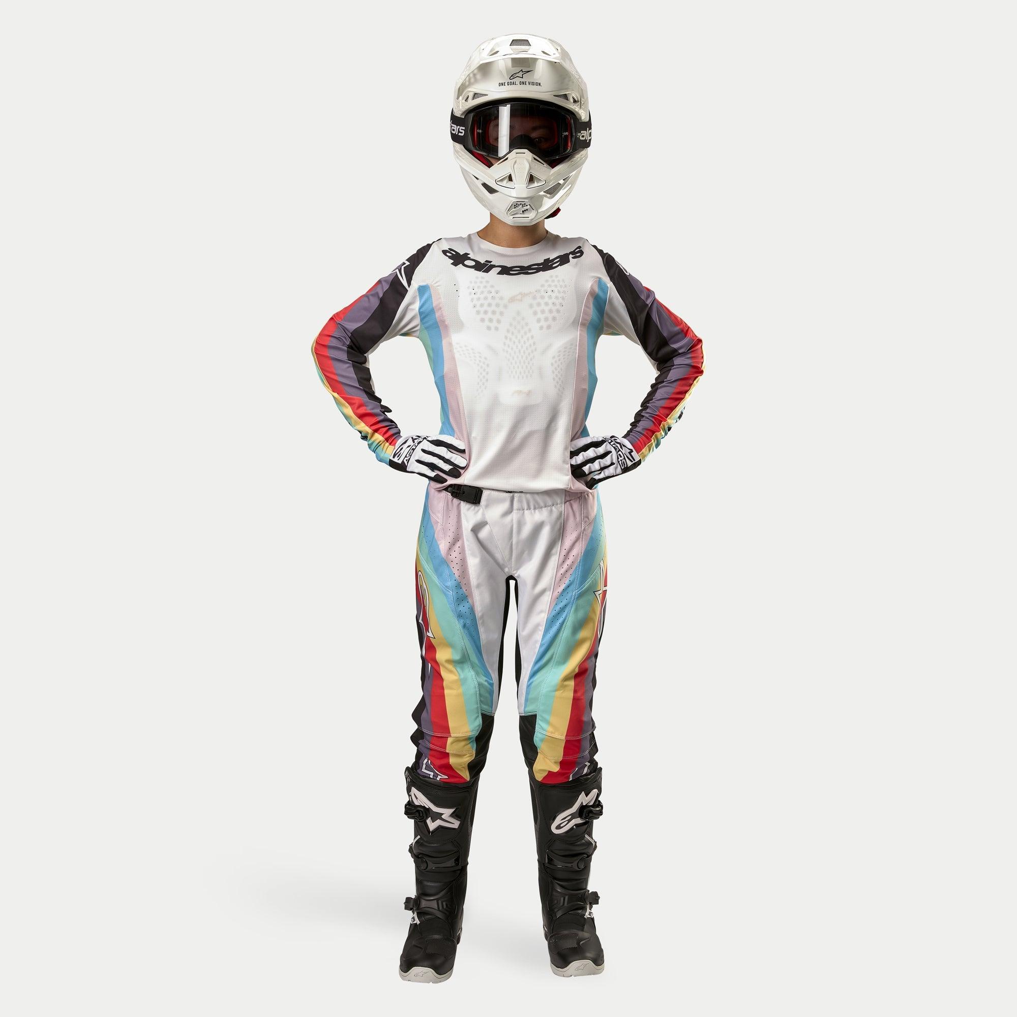 Kvindelig motocross-kører iført Alpinestars Stella 2024 Techstar bukser til damer, Black Multicolor med hvid base samt røde, gule og blå striber, matchende langærmet trøje, hvid hjelm og sorte støvler, komplet racerudstyr til off-road konkurrence