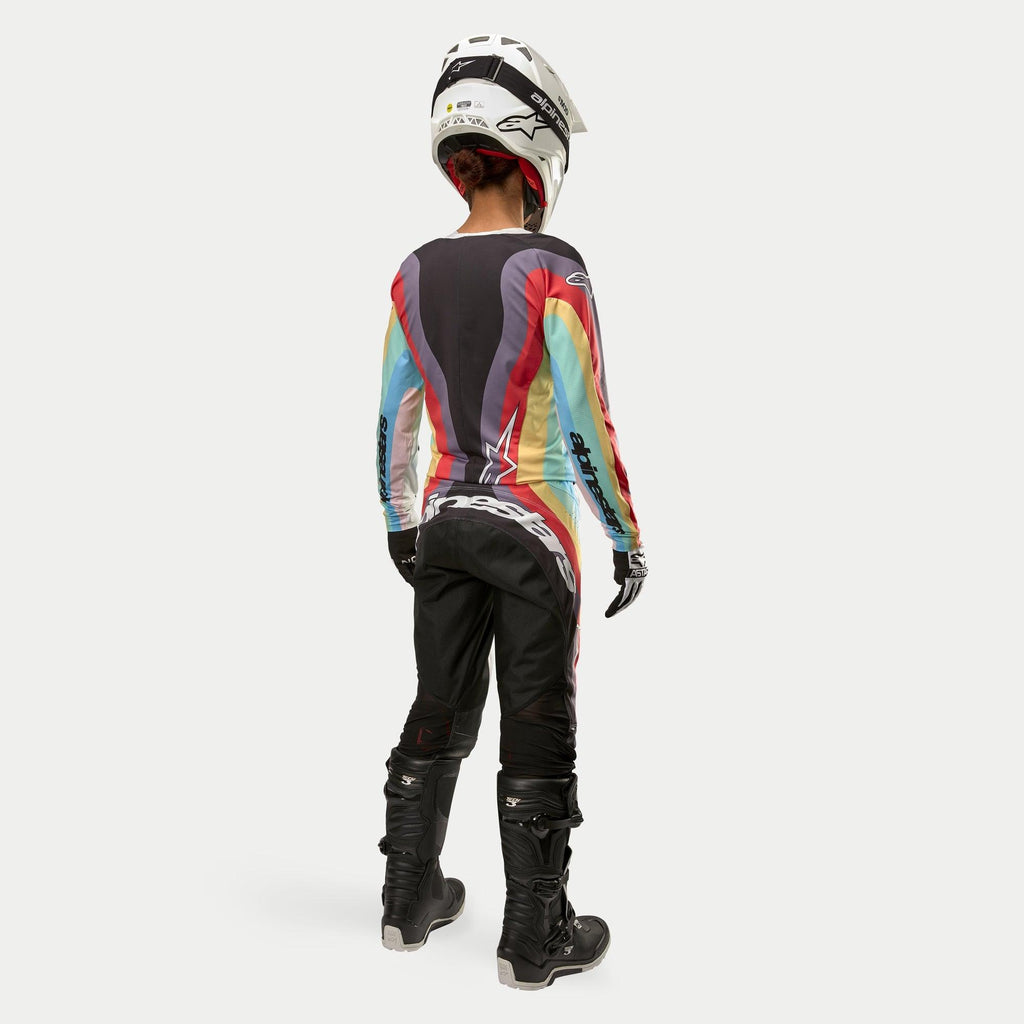 Motocrosskører set bagfra iført Alpinestars Stella 2024 Techstar bukser til kvinder i Black Multicolor, med stort hvidt logo på sædet, koordineret med en farverig stribet jersey, hvid hjelm og sorte Tech 3 støvler til off-road racing.