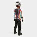 Motocrosskører set bagfra iført Alpinestars Stella 2024 Techstar bukser til kvinder i Black Multicolor, med stort hvidt logo på sædet, koordineret med en farverig stribet jersey, hvid hjelm og sorte Tech 3 støvler til off-road racing.