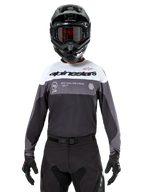 Motocross-rytter iført Alpinestars Pro-Dura Jersey i Black Magnet White, langærmet performance-trøje med hvide og grå farveblokke, bæres med sort hjelm og goggles, med 'alpinestars' branding og 'ONE GOAL ONE VISION' slogan, off-road motorcykel-racingudstyr