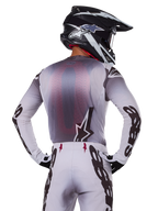 Alpinestars 2025 Supertech Maker udstyrssæt, motocross-rytter set bagfra, hvid og grå jersey med stor sort branding på ærmerne og stjernelogo, matchende hvide bukser med sort grafik, sort og hvid hjelm, professionelt terrænkørselsudstyr