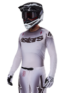 Alpinestars 2025 Supertech Maker-udstyrssæt i hvid, motocross-kører med en grå gradueret jersey, sort astars-logo og laserskårne perforeringer, matchende bukser med magnetisk spænde og stjernegrafik, sort og hvid hjelm og handsker, komplet off-road racing-look.