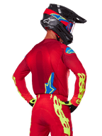 Bagfra af en motocrosskører iført Alpinestars 2025 Supertech Maker-udstyrssæt i rød, bestående af en rød jersey med neongul branding på ærmerne, matchende røde bukser, en sort hjelm med blå og røde detaljer samt koordinerende sorte og gule handsker.