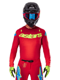 Alpinestars 2025 Supertech Maker Jersey i Bright Red Yellow Fluo, med gult 'astars' brystlogo og blå stjernedesigns på skuldrene, båret med matchende bukser, sort hjelm, blå goggles og gule handsker, professionelt motocross-racerudstyr