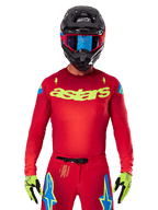 Alpinestars 2025 Supertech Maker Jersey i Bright Red Yellow Fluo, med gult 'astars' brystlogo og blå stjernedesigns på skuldrene, båret med matchende bukser, sort hjelm, blå goggles og gule handsker, professionelt motocross-racerudstyr