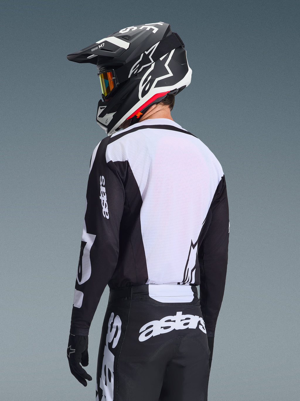 Motocrosskører set bagfra iført Alpinestars 2026 Racer Air Riway-trøje, sort og hvid, med hvidt åndbart mesh-rygpanel og sorte ærmer med 'astars'-branding, matchende sorte bukser med hvide logoer, sort hjelm med hvide detaljer, professionelt off-road udstyr til præstation i høje temperaturer.