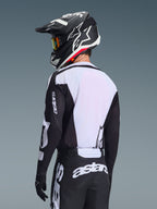 Motocrosskører set bagfra iført Alpinestars 2026 Racer Air Riway-trøje, sort og hvid, med hvidt åndbart mesh-rygpanel og sorte ærmer med 'astars'-branding, matchende sorte bukser med hvide logoer, sort hjelm med hvide detaljer, professionelt off-road udstyr til præstation i høje temperaturer.