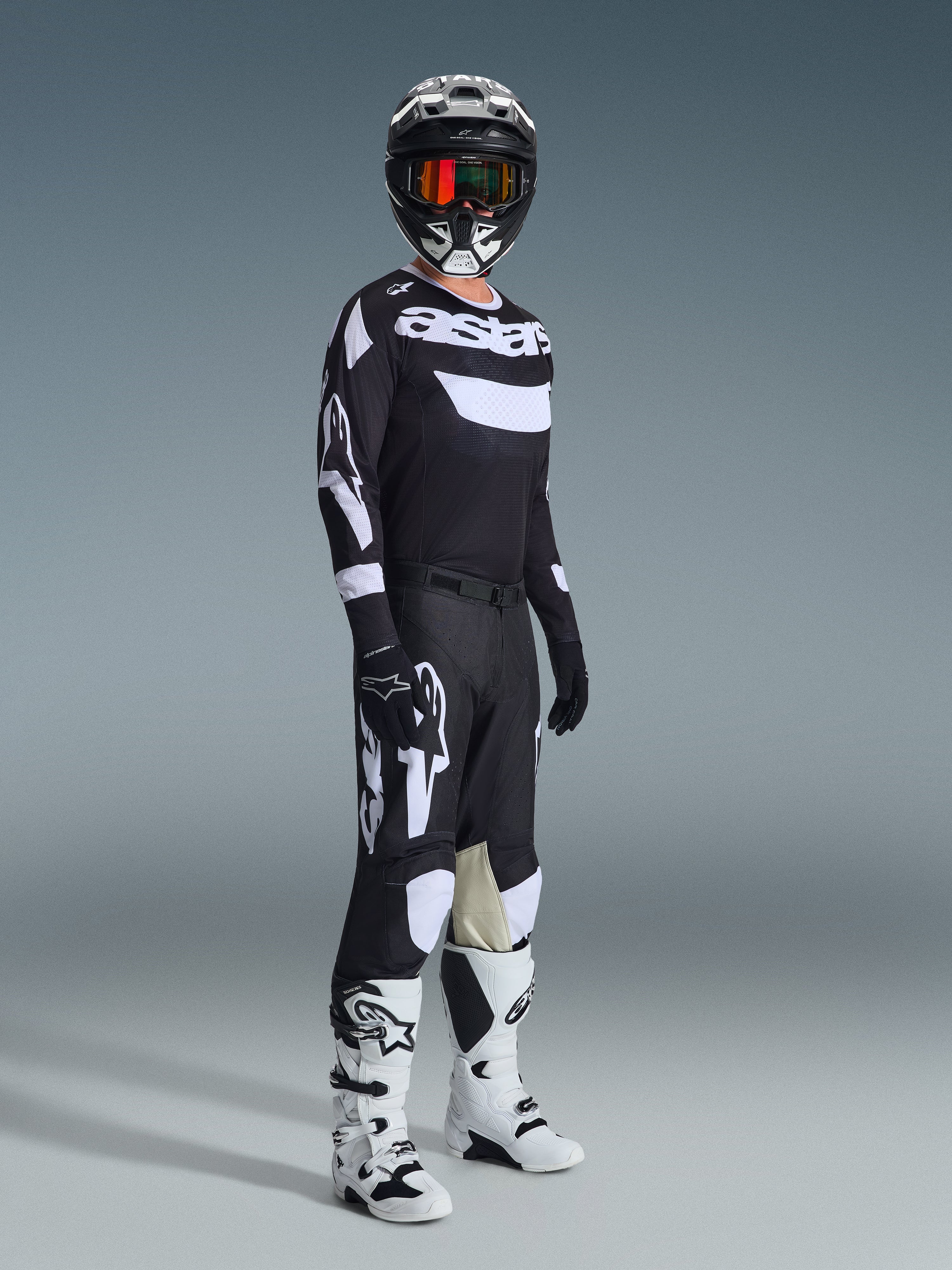 Motocrosskører i komplet racerudstyr, 2026 Racer Air Riway-trøje i sort og hvid, matchende sorte bukser med store hvide Alpinestars-logoer, sort hjelm med hvide detaljer og orange goggles, hvide og sorte støvler, koordineret outfit til professionel off-road kørsel