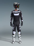 Motocross-kører iført komplet racerudstyr, Alpinestars 2026 Racer Air Riway-trøje i sort og hvid med 'astars'-branding på brystet, matchende sorte bukser med hvide detaljer, hvide motocross-støvler og sort og hvid hjelm med goggles, koordineret professionelt off-road raceroutfit