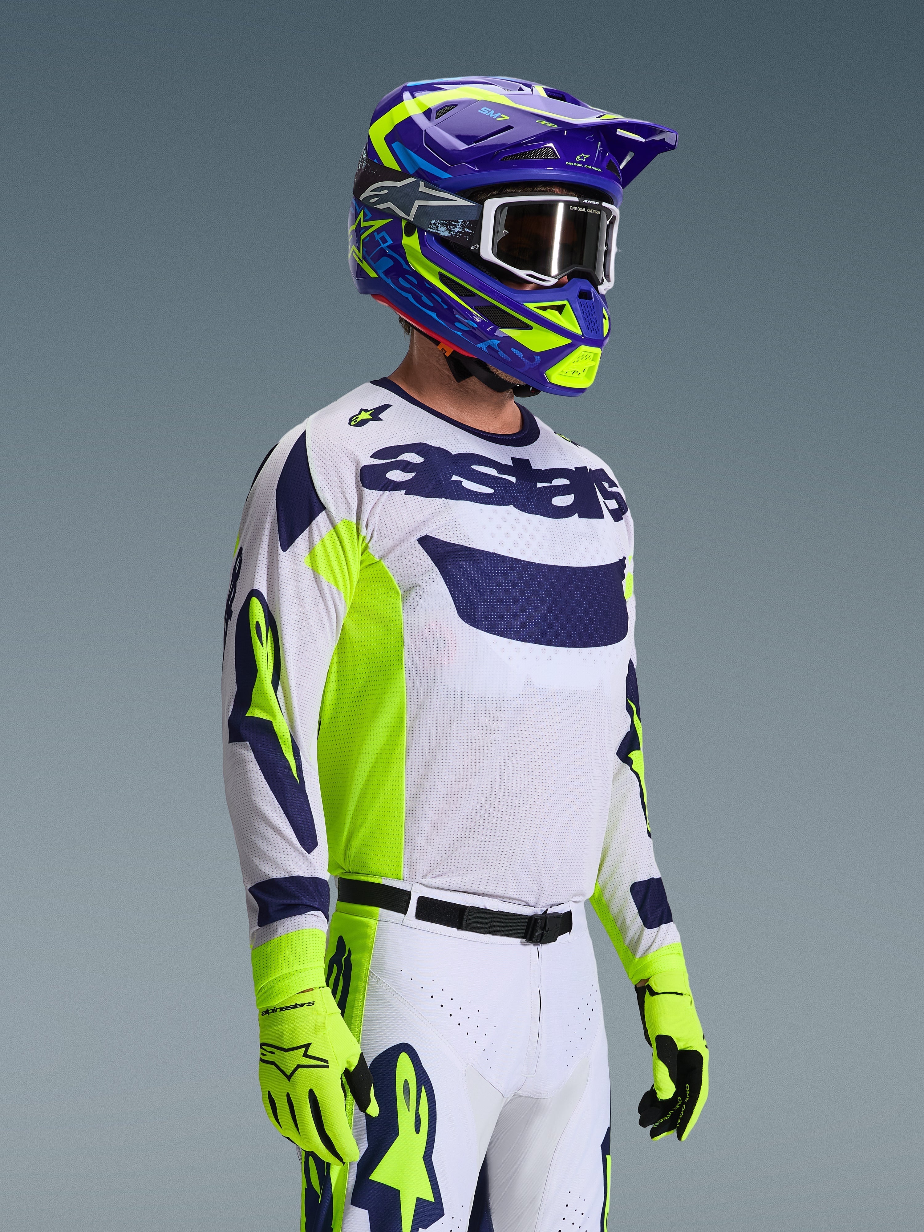 Motocross-rytter iført Alpinestars 2026 Racer Air Riway-trøje, lysegrå med fluo-gule og blå detaljer, kombineret med en matchende blå og gul hjelm og fluo-gule handsker, med åndbar mesh-konstruktion og professionel racer-styling til off-road ydeevne.