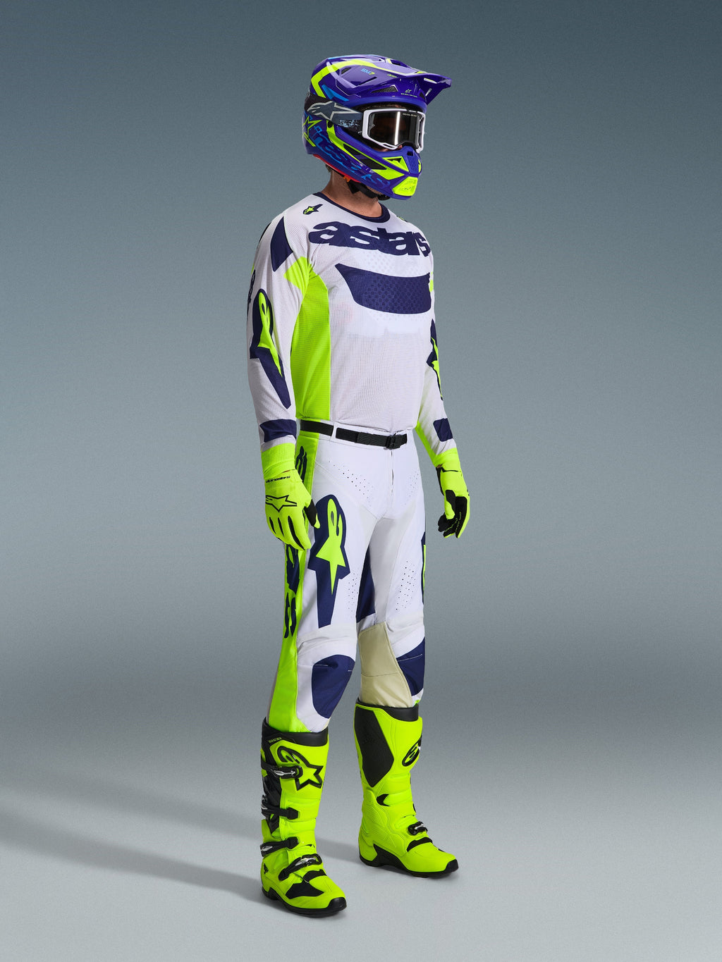 Alpinestars motocross-kører i komplet racerudstyr, lysegrå trøje med Yellow Fluo og blå detaljer, matchende hvide og gule bukser, neongule støvler og handsker, lilla og gul hjelm med mørke briller, koordineret outfit til off-road kørsel