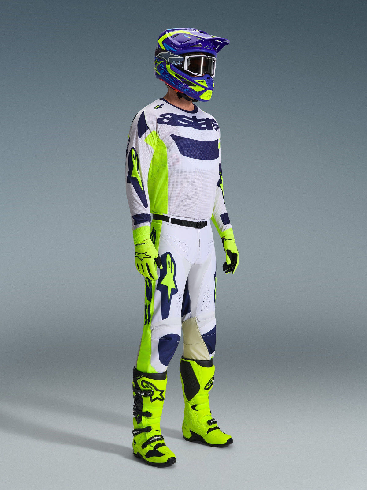Motocross-kører iført Alpinestars 2026 Racer Air Riway-udstyrssæt i lysegrå, neongul og blå. Sættet inkluderer en langærmet mesh-trøje, matchende racerbukser, klare neongule handsker og støvler samt en lilla og blå hjelm med goggles, designet til ventileret off-road ydeevne.