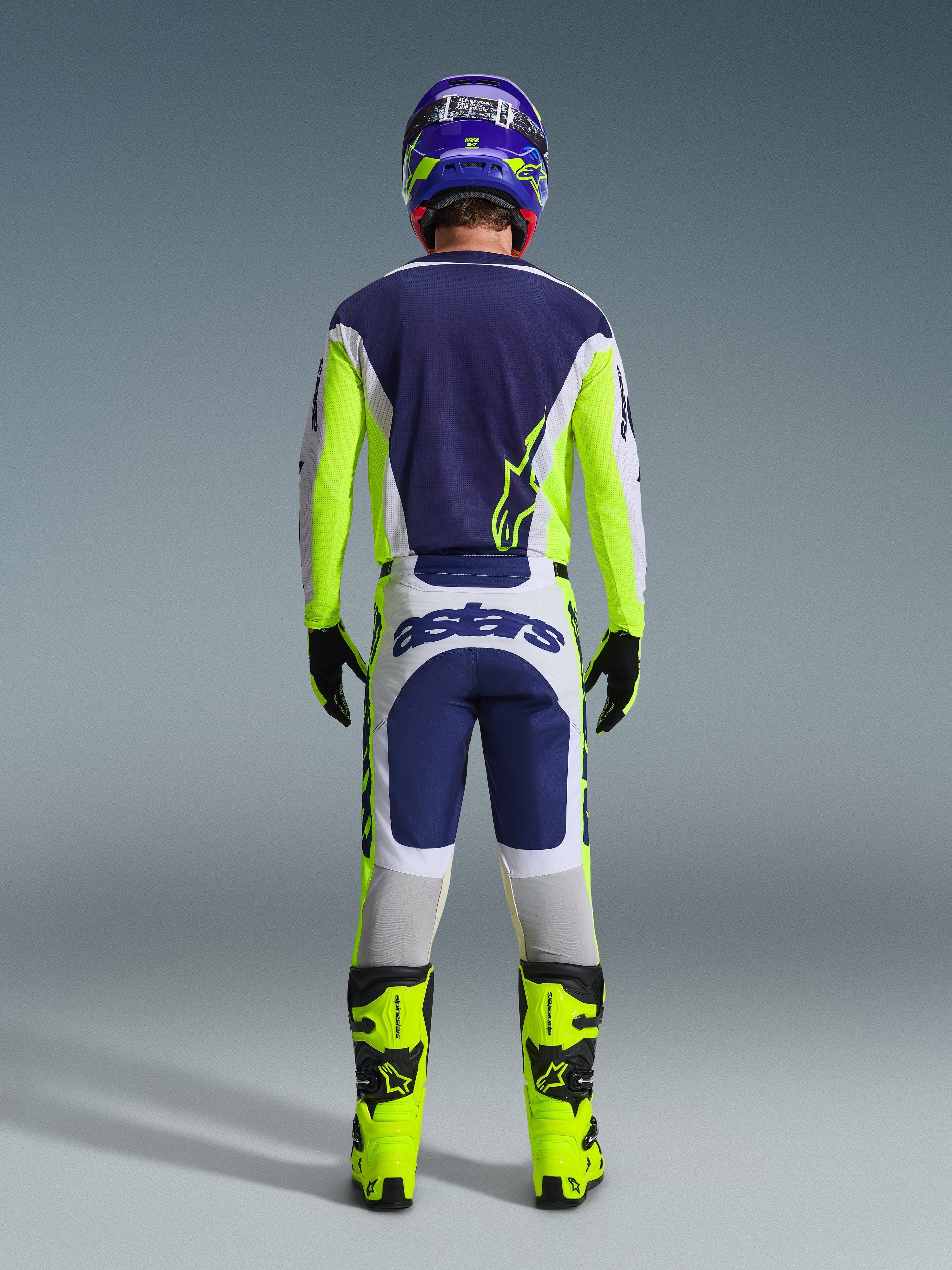 Bagsidevisning af en motocrosskører iført Alpinestars 2026 Racer Air Riway-trøje og matchende bukser i Light Gray Yellow Fluo Blue, kombineret med neongule støvler og en matchende hjelm, som viser et komplet off-road racersæt.