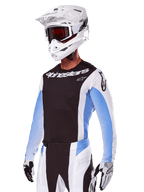 Motocrossrytter iført Alpinestars 2025 Techstar Melt udstyrssæt, hvid med sorte og lyseblå detaljer, bestående af en jersey med sort bryst og hvide ærmer, matchende hvide og sorte bukser, hvid SM10-hjelm med goggles og sort-hvide handsker, komplet professionelt off-road raceroutfit.