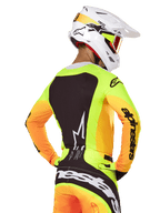 Motocross rider wearing Alpinestars 2025 Techstar Melt Jersey, back view, neon yellow with orange sleeves and a large black graphic panel, matching motocross pants with bold branding, white Alpinestars helmet and orange goggles, professional off-road racing gear