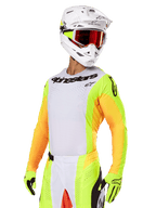 Motocross-kører iført Alpinestars 2025 Techstar Melt-udstyrssæt, hvid jersey med livlige gule og orange ærmer, sort 'Alpinestars' branding på brystet, matchende neongule handsker, hvid hjelm med rødfarvede briller, professionelt off-road racerudstyr
