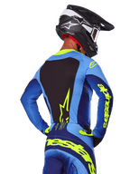Rear view of a motocross rider wearing the Alpinestars 2025 Techstar Melt Jersey in Blue and Yellow Fluo, featuring a black back panel and bold yellow branding on the sleeves, paired with matching motocross pants and a black carbon helmet, performance off-road racing gear