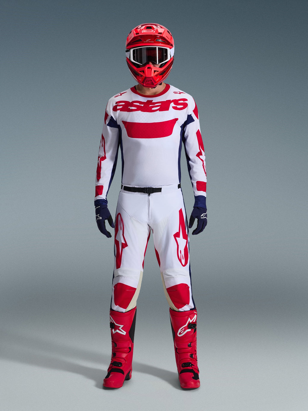 Motocross-kører iført komplet racerudstyr, Alpinestars 2026 Racer Riway jersey i hvid, blå og rød, matchende hvide og røde bukser, rød hjelm, røde støvler og mørkeblå handsker, koordineret outfit til off-road kørsel