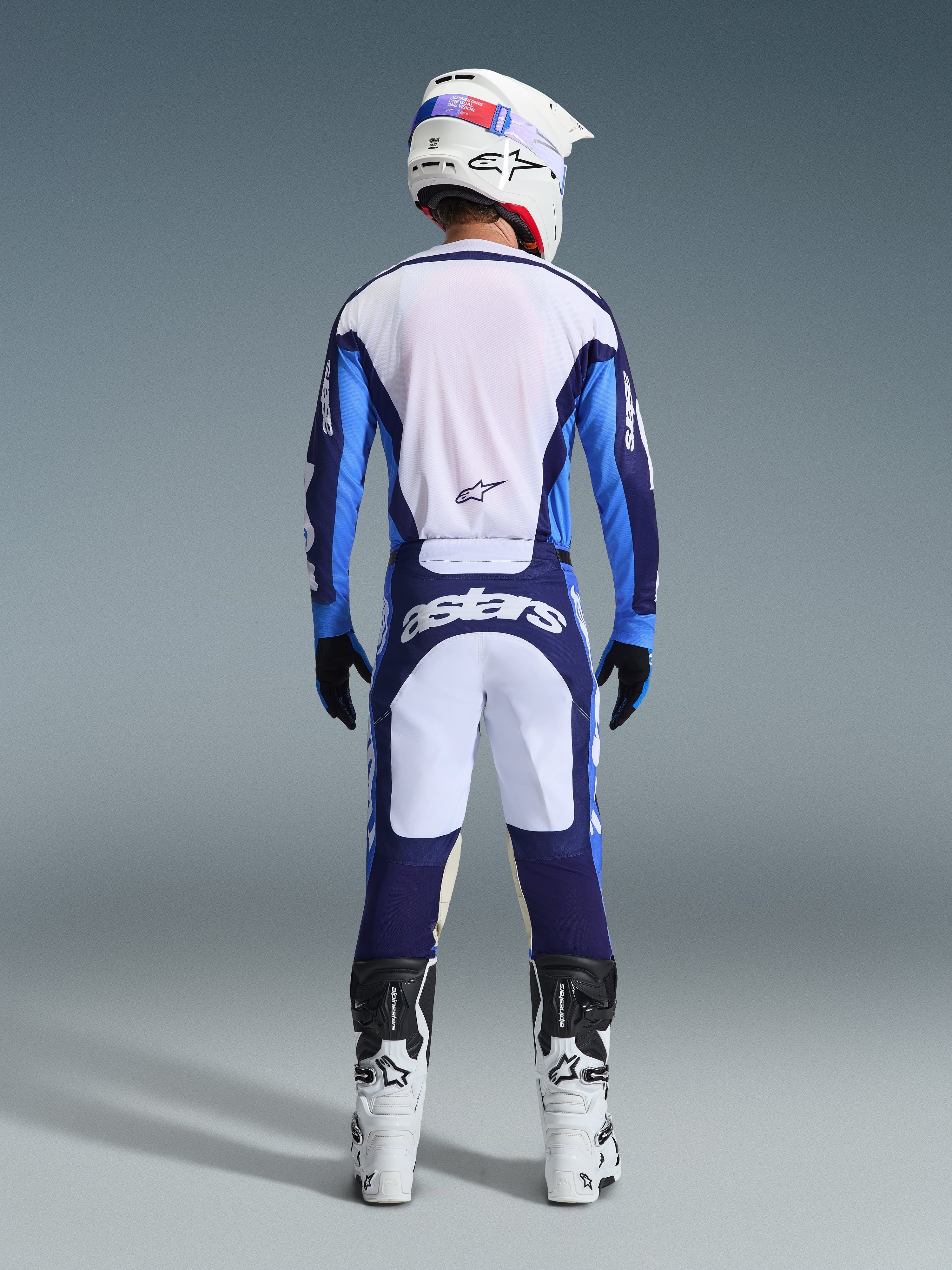 Set bagfra af en motocrosskører iført Alpinestars 2026 Racer Riway Jersey og bukser i Ucla Blue og hvid, koordineret med en hvid hjelm, sorte handsker og hvide motocrossstøvler, højpræstationsudstyr til off-road racing
