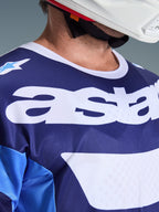 Nærbillede af Alpinestars 2026 Racer Riway trøje i Ucla Blue og hvid, med et stort hvidt 'astars'-logo på brystet, hvid krave og åndbar mesh-tekstur, båret med en hvid motocross-hjelm